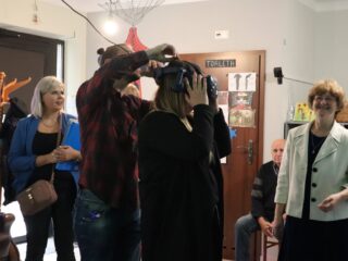 Mężczyzna zakłada kobiecie okulary VR. Wokół stoi kilka uśmiechniętych osób.