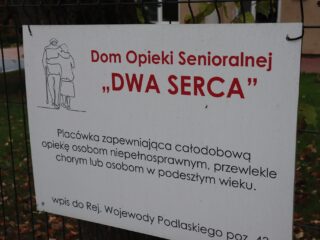 Tabliczka Domu Opieki Senioralnej DWA SERCA zawieszona na siatce.