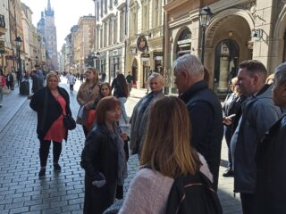 Grupa osób stoi i rozmawia między sobą na ulicy przechodzącej między łączonymi zabytkowymi budynkami miasta.