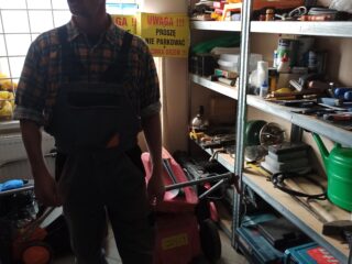 Mężczyzna w roboczych ogrodniczkach stoi w pomieszczeniu ze sprzętem. Po prawej stronie stoi półka z narzędziami.