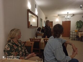 Grupa kobiet siedzi przy stolikach które stoją przy ścianie w jednym rzędzie. Dwie kobiety patrzą w stronę kobiety która stoi przy stoliku i przemawia.