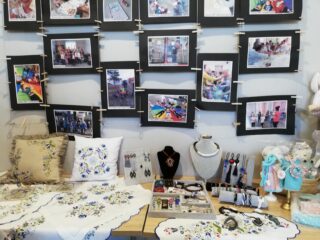 Stół z biżuterią, poduszkami i serwetkami w kaszubskie wzory. Nad stołem na ścianie wiszą zdjęcia.