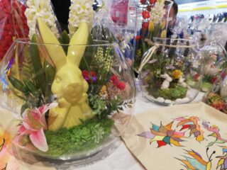 Ozdoby wielkanocne m.in. materiałowe torby, kwiaty oraz ozdoby z królikami w okrągłych szklanych dzbankach.