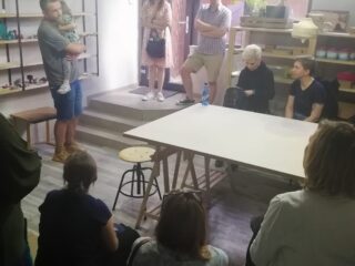 Grupa osób siedzi w pomieszczeniu w którym na środku stoi pusty stół. Dwie osoby stoją przy drzwiach. Jeden z stojących mężczyzn trzyma dziecko.