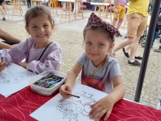 11 Podlaskie Targi Ekonomii Społecznej w Białymstoku na Rynku Kościuszki. Dwie uśmiechnięte dziewczynki siedzą przy stole i kolorują kolorowanki. W tle widać wystawę fotograficzną.