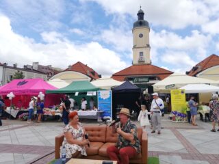 11 Podlaskie Targi Ekonomii Społecznej w Białymstoku na Rynku Kościuszki. Dwie osoby z mikrofonami siedzą na kanapie na tle rozstawionych stoisk i ratusza.