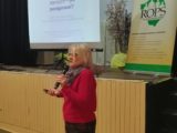Kobieta w czerwonym swetrze stoi przy scenie i przemawia przez mikrofon. Na scenie stoi ekran projekcyjny z wyświetloną prezentacją.