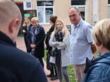 Kilka osób stoi przed wejściem do budynku na chodniku z kostki i patrz w stronę jednej z osób.
