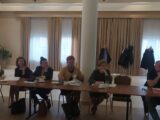 Kilka osób siedzi przy długim stole na którym leżą dokumenty. Wszyscy patrzą w jednym kierunku.