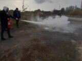 Osoba przy użyciu gaśnicy gasi ognisko. Obok stoi kilka osób. Wydarzenie odbywa się na świeżym powietrzu.