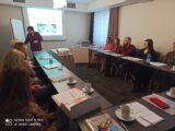 Warsztaty na temat pracy z osobami niepełnosprawnymi intelektualnie. Grupa osób siedzi przy stołach ustawionych w kształt litery U i patrzy na kobietę prowadzącą która notuje na tablicy.