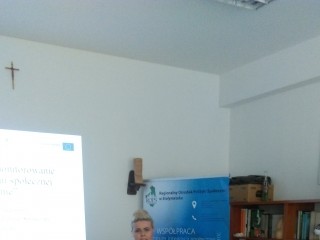 Kobieta która trzyma dokument stoi przed stołem z projektorem i poczęstunkiem. Za nią na ścianie jest wyświetlona prezentacja.