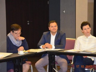 Trzy osoby siedzą na rogu łączonych stołów na których leżą teczki z dokumentami.