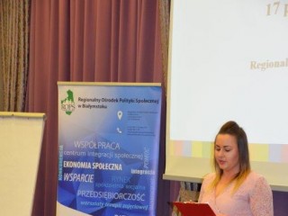 Kobieta stoi na tle ekranu projekcyjnego z wyświetloną prezentacją i czyta dokument. Obok stoi baner.