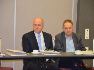 Dwóch mężczyzn w marynarkach siedzi przy jednym stole obok siebie. Na stole leżą teczki z dokumentami.