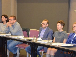 Kilka osób siedzi przy jednym stole i patrzy w jednym kierunku. Na stole leżą teczki.