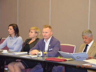 Cztery osoby siedzą blisko siebie przy jednym stole na którym znajdują się teczki. Jeden z mężczyzn przegląda teczkę.