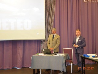 Dwóch mężczyzn w garniturach stoi przy stole z laptopem obok wiszącego ekranu projekcyjnego z wyświetloną prezentacją.