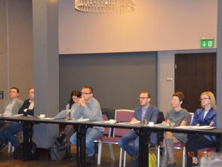 Grupa osób siedzi przy stole na dużej sali i patrzy w jednym kierunku. Na stole leżą teczki. Z tyłu widać wyjście ewakuacyjne.