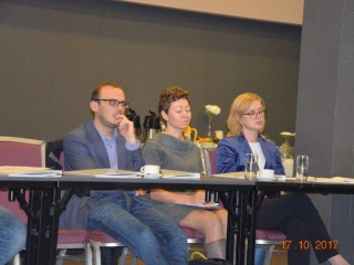 Trzy zamyślone osoby siedzą obok siebie przy jednym stole. Na stole leżą teczki z dokumentami.