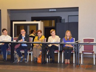 Pięć osób siedzi przy łączonych stołach i patrzy w jednym kierunku. Na stołach leżą teczki i dokumenty.