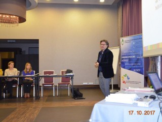 Kobieta stoi na tle ekranu projekcyjnego z wyświetloną prezentacją i przemawia do publiczności która siedzi przy stole.