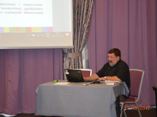 Mężczyzna siedzi przy stoliku z laptopem obok ekranu projekcyjnego który wisi z wyświetloną prezentacją.