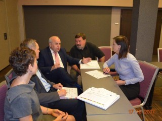 Kilka osób siedzi na rogu łączonych stołów i rozmawia między sobą. Na stole leżą dokumenty.