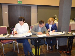 Trzy osoby siedzą blisko siebie przy długim stole i podpisują dokumenty. Z tyłu widać wyjście ewakuacyjne.