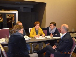 Pięć osób siedzi blisko siebie przy jednym długim stole na którym leżą dokumenty i rozmawiają między sobą.