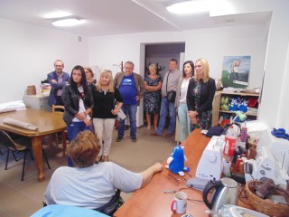 Grupa osób stoi przy wejściu do pracowni w której siedzi kobieta przy stole z maszynami do szycia i pluszowymi zabawkami.