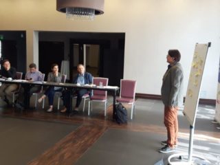 Osoba prowadząca stoi przy tablicy przed resztą uczestników spotkania którzy siedzą przy łączonych stołach.