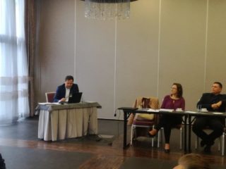 Osoba prowadząca siedzi przy laptopie a obok przy drugim stole siedzi reszta uczestników spotkania.