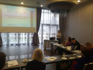 Grupa osób siedzi przy stole na przeciwko ekranu prezentacyjnego.