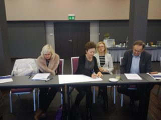 Cztery osoby siedzą przy jednym stole i pracują nad projektem. W tle widać drzwi wyjściowe.