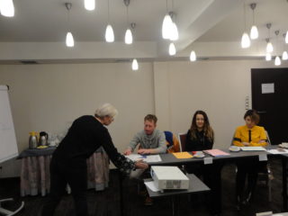 Trzy osoby siedzą przy stole z dokumentami. Obok stoi osoba prowadząca która sięga w stronę projektora.