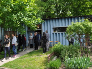 Grupa osób wychodzi z niewielkiego, niebieskiego budynku z ogródkiem. Kilka osób rozmawia przy wejściu.