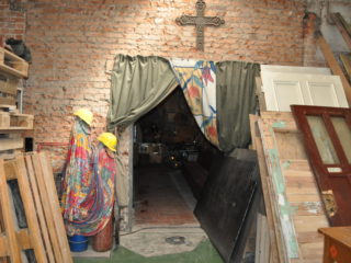 Stare pomieszczenie ze ścianami z czerwonej cegły i krzyżem nad wyjściem. W środku znajdują się m.in. drewniane drzwi, palety, płachty i deski.