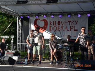 Zespół muzyczny z pełnym sprzętem muzycznym stoi na scenie i gra. Z tyłu wisi baner Kilometry Dobra.