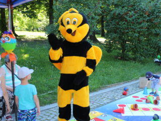 Osoba w stroju pszczoły na tle zieleni oraz dzieci które się grają w gry planszowe.