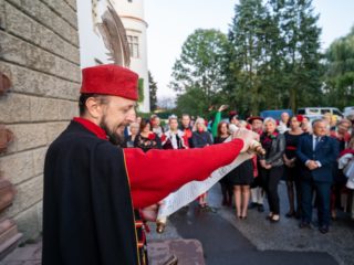 Mężczyzna w czerwono-czarnym stroju w czapce z piórem stoi przy budynku przed publicznością i czyta ze zwoju.