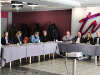 Grupa osób siedzi przy łączonych stołach. Na środku sali stoi projektora a z tyłu za uczestnikami widać wieszaki z kurtkami.