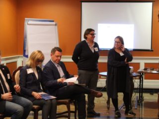 Trzy osoby siedzą na krzesłach a obok stoją dwie uśmiechnięte kobiety przy których stoi projektor. Z tyłu widać ekran projekcyjny i tablicę.