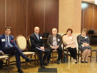 Kilka osób siedzi w jednym rzędzie na krzesłach przy ścianie. Wszyscy patrzą w jedną stronę.