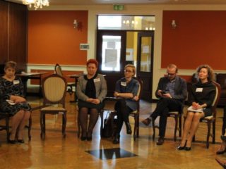 Kilka osób siedzi na krzesłach w jednym rzędzie. Z tyłu widać drzwi wyjściowe.