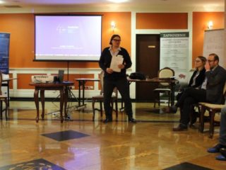 Kobieta prowadząca spotkanie stoi obok projektora i przemawia do osób które siedzą z boku w jednym rzędzie na krzesłach. W tle widać ekran projekcyjny z wyświetloną prezentacją.