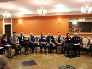 Grupa osób siedzi na krzesłach w półokręgu w sali z pomarańczowymi ścianami, marmurową podłogą i kryształowymi żyrandolami.