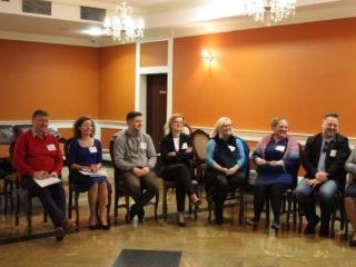 Grupa uśmiechniętych osób siedzi na krzesłach w półokręgu w sali z pomarańczowymi ścianami, marmurową podłogą i kryształowymi żyrandolami.