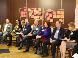 Kilka osób siedzi na krzesłach w jednym rzędzie przy ścianie na której wiszą kolorowe kartki z notatkami.