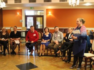 Kobieta z teczką w rękach stoi i przemawia do uczestników spotkania którzy siedzą na krzesłach wokół. W tle widać drzwi wyjściowe.